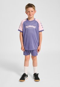 Lila Sportoutfit mit pinkfarbenen Akzenten, das vertikale Streifen und einen Rundhalsausschnitt aufweist, kombiniert mit passenden Shorts und schwarzen Sneakers.