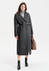 Szary płaszcz w jodełkę z paskiem, z klapami, bocznymi kieszeniami, do kolan, zestawiony z szarym golfem i czarnymi botkami za kostkę.