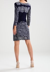Navy-Kleid mit langen Ärmeln, das komplizierte weiße Spitzenmuster aufweist. Ein tailliertes Design mit rundem Ausschnitt und einfarbigen Akzenten.