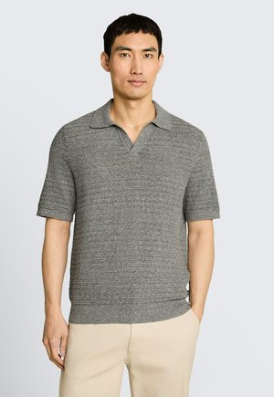 Homme portant un polo tricoté gris à manches courtes et un pantalon beige, debout devant un fond clair uni.