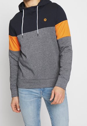 Mężczyzna noszący bluzę z kapturem w stylu color-block z granatową górą, pomarańczowymi paskami na rękawach i szarym korpusem, w połączeniu z jasnoniebieskimi dżinsami.