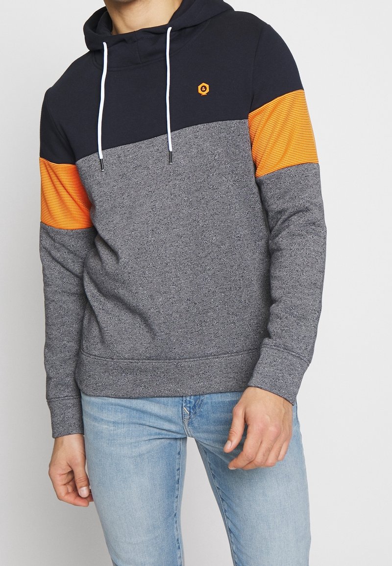 Homme portant un sweat à capuche color block avec le haut bleu marine, des bandes orange sur les manches et le corps gris, associé à un jean bleu clair.