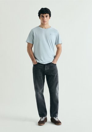 Joven de pie con las manos en los bolsillos, llevando una camiseta azul clara, vaqueros negros y zapatillas marrones frente a un fondo claro y liso.