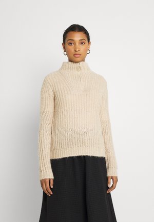 Beige knitted sweater with a high collar and zipper, featuring a ribbed texture. Paired with a black textured skirt.