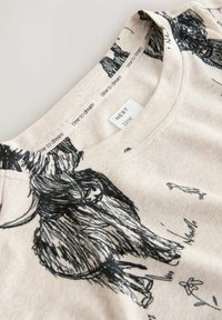 Camiseta beige claro con cuello redondo, con ilustraciones negras de animales y plantas, confeccionada en algodón suave. Etiquetas visibles en el interior del cuello.