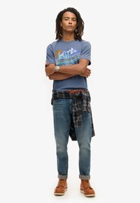 Blå grafisk t-shirt med et natursmotiv, iført med lyseblå jeans og brune snørebåndsstøvler. En sort ternet skjorte er bundet om taljen.