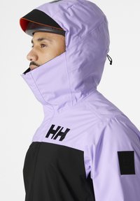 Lavendel en zwarte waterproof jas met hoge kraag, verstelbare capuchon en logodetail. Textuurstof en slank ontwerp zichtbaar.