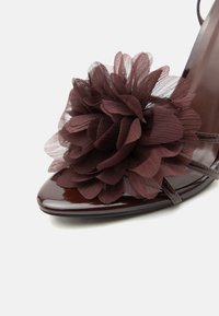 Λαμπερό σκούρο καφέ παπούτσι με ψηλό τακούνι, διακοσμημένο στην μύτη με μεγάλο στρωματοποιημένο υφασμάτινο λουλούδι και λεπτά δερμάτινα λουράκια.