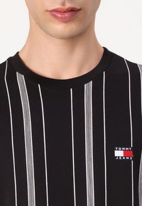 Close-up of a person wearing a black shirt with vertical white stripes and a small Tommy Jeans logo patch on the chest.