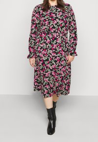 Frau, die ein schwarzes Midikleid mit rosa Blumenmuster und langen Ärmeln trägt, kombiniert mit schwarzen Stiefeletten, die vor einem einfarbigen Hintergrund läuft.