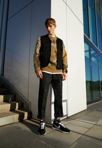 Jeune homme en pull camouflage, gilet noir, pantalon noir retroussé et baskets Nike noires, debout à l'extérieur près d'un bâtiment moderne et des escaliers.