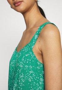 Grüner, blumengemusterter ärmelloser Top mit zarten weißen Blumen, einem gewellten Ausschnitt und verstellbaren Trägern, aus leichtem Stoff gefertigt.