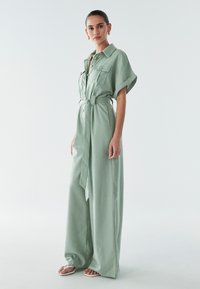 Svetlo zelena lanena kombinezon s ovratnikom, kratkim rukavima, žepovima, pojasom u struku i širokim hlačama. Glatka tekstura i opušten kroj.