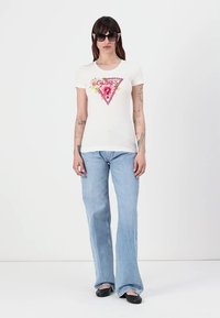 Witte katoenen t-shirt met een roze driehoekig logo met bloemaccenten. Gecombineerd met lichtblauwe flared jeans en zwarte platte schoenen.