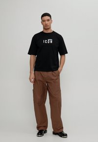 Sort t-shirt med hvid "ICON" tryk, parret med brune cargo bukser og sorte sko. Enkel, afslappet pasform, minimalistisk design.