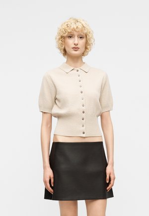 Žena s krátkymi kučeravými blond vlasmi, oblečená v béžovom pletenom svetri na gombíky s krátkym rukávom a čiernej koženej mini sukni, stojaca na bielom pozadí.