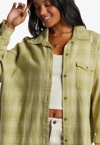 Grøn- og hvidternet skjorte med knapstolpe og lomme, lagdelt over en ribbet hvid top, boret med lyse bukser.