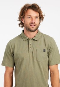 Olijfgroene polo met een kraag, drie knopen en een borstzak. Heeft een textuurstof en een klein logo-embleem.