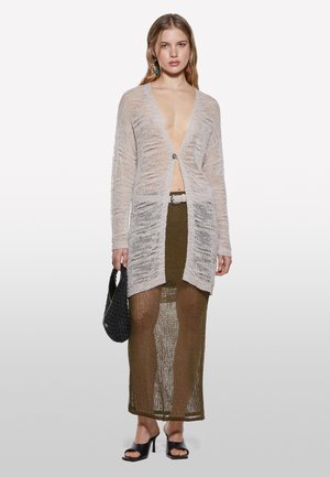 Leichte, offen gestrickte Strickjacke in sanftem Beige mit langen Ärmeln und einer Frontknopfleiste, kombiniert mit einem grünen Netz-Maxikleid. Schwarze Handtasche.
