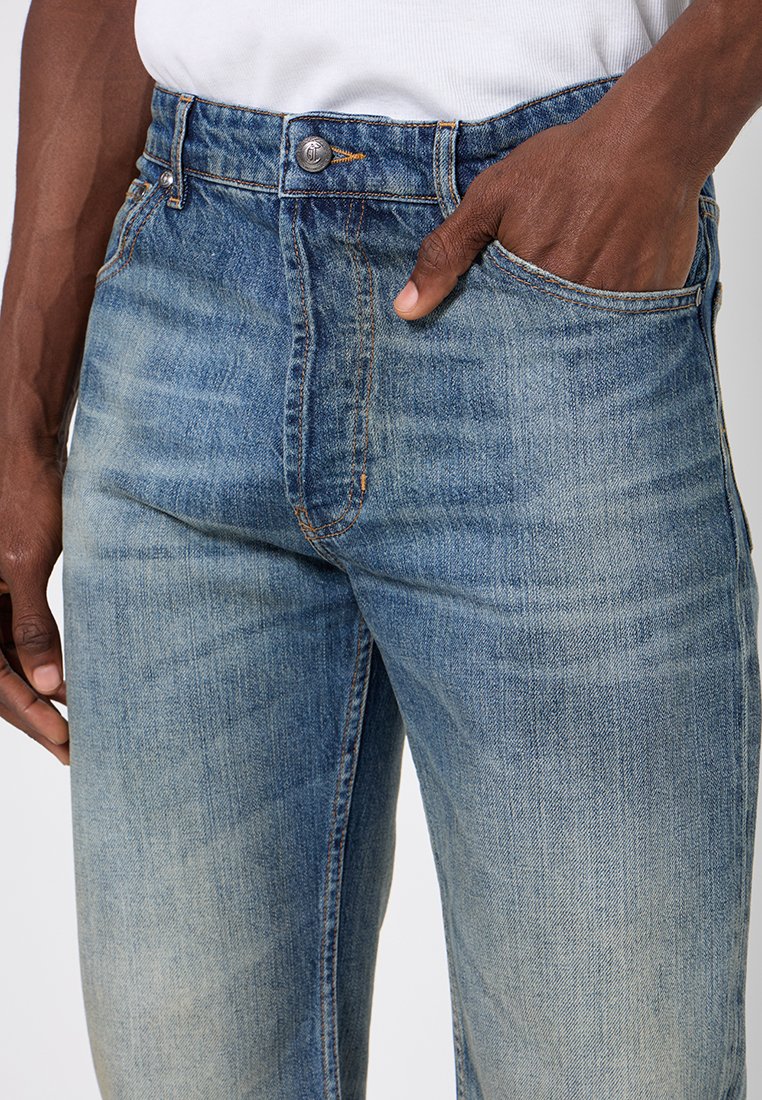 Jeans en denim bleu clair avec un design classique à cinq poches, une fermeture à bouton en métal et des détails de décoloration subtils. Tissu texturé avec des accents de couture.