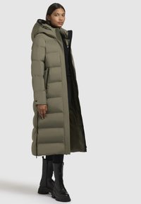 Lang, gevoerd jasje in olijfgroen, met een capuchon, een rits aan de voorkant en zijzakken. Gekleed over een zwart top en gecombineerd met zwarte veterschoenen.