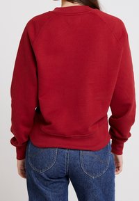Red sweatshirt with a ribbed collar, dropped shoulder seams, and a soft texture. Paired with blue jeans featuring back pockets.