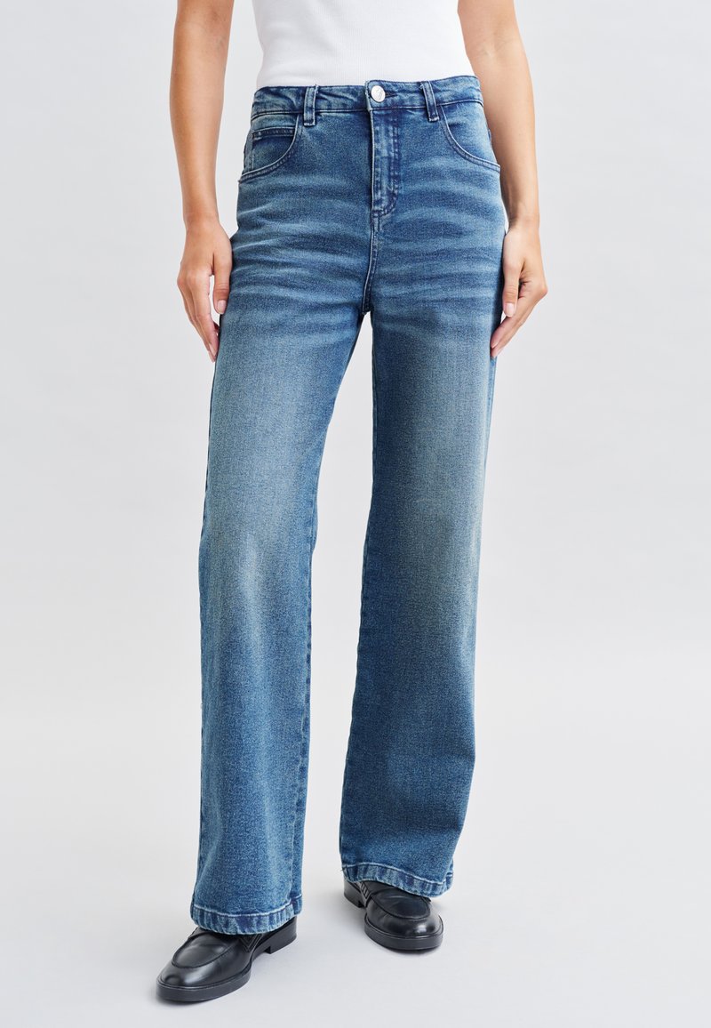 Jean bleu à jambes larges en denim, avec une taille haute, une fermeture à bouton, et un délavage subtil. Associé à des chaussures noires.