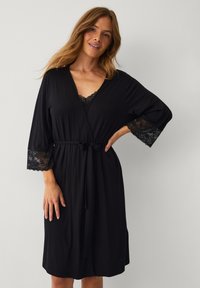 Robe noire en tissu doux, ornée de détails en dentelle sur les manches, avec une ceinture à nouer, un ajustement décontracté et un décolleté en V.