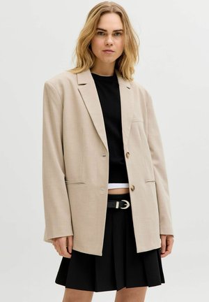 Junge Frau mit blondem Haar trägt einen beigen oversized Blazer, ein schwarzes kurzes Top, einen schwarzen Faltenrock und einen schwarzen Gürtel mit silberner Schnalle.