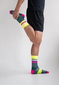 Bunte Sport-Socken mit einem dunkelgrünen Grundton, pinken und neongelben Streifen sowie einer Kombination aus verschiedenen Texturen. Das Design umfasst einen auffälligen Akzent.