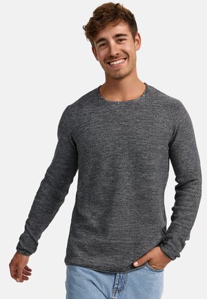 Ung mand, der smiler, iført en mørkegrå tekstureret langærmet trøje og lyseblå jeans, står med den ene hånd i lommen.