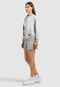 Woman wearing light grey bomber jacket, white t-shirt, distressed denim shorts, and white high-top sneakers, standing sideways on gray background.