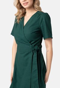 Zelené šaty ve stylu wrap z texturovaného materiálu, s krátkými rukávy, výstřihem do V, bočním zavázáním a splývavou sukní s jemným drapáním.