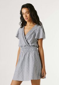 Jauna moteris stovinti, dėvinti trumpomis rankovėmis mėlynai baltą dryžuotą suknelę, surištą juosmeniu, su subtiliu gėlių raštu.