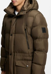 Braune gepolsterte Jacke mit Kapuze, ausgestattet mit einer Reißverschlusstasche an der Seite, Druckknöpfen und texturiertem Futter. Einfaches schwarzes Logo-Patch auf der Schulter.