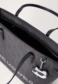 Bolso tote negro con exterior texturizado, patrón en relieve, asas negras, cierre superior con cremallera y un llavero de gato de dibujos animados como acento.