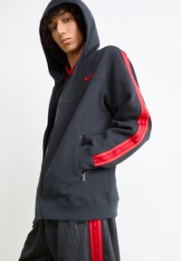 Hoodie preto com fecho de correr, feito de fleece, com um logótipo vermelho da Nike no peito e riscas vermelhas em contraste nas mangas. Inclui bolsos laterais.