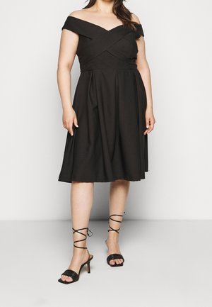 Kobieta w czarnej sukience za kolano, z odkrytymi ramionami i plisowanym dołem, oraz w czarnych sandałach na wysokim obcasie z paskami, na jednolitym tle.