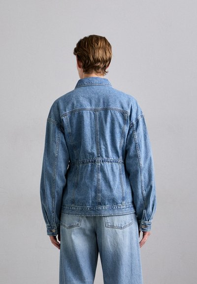 Denims jaka ar gaiši zilu toni, ar izsistām vidukli, garām piedurknēm un klasisku apkakli. Redzami šuvju sīki elementi uz muguras.