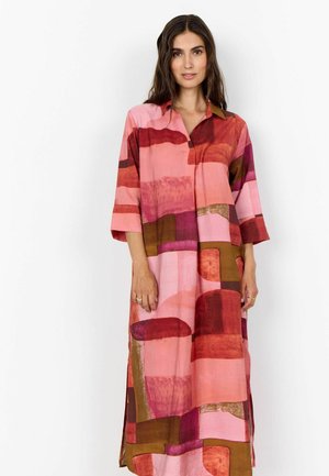 Žena oblečená v dlhých, voľne padnúcich šatách s abstraktnými vzormi v ružovej, červenej a hnedej farbe, stojaca na bielom pozadí.
