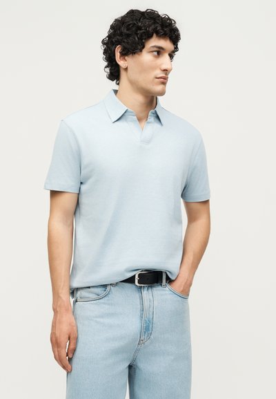 Νέος άνδρας με σγουρά σκούρα μαλλιά, φοράει ανοιχτό μπλε πόλο με κοντά μανίκια και ανοιχτό μπλε τζιν, στέκεται με το ένα χέρι στην τσέπη.