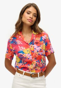 Chemise à manches courtes de couleur rose vif avec des motifs floraux en jaune, bleu et blanc. Présente une poche sur la poitrine et des détails de boutons.