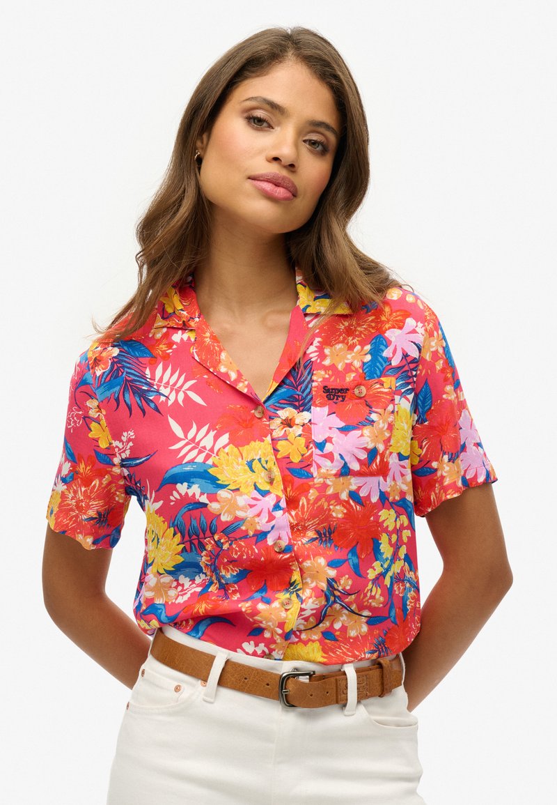 Chemise à manches courtes de couleur rose vif avec des motifs floraux en jaune, bleu et blanc. Présente une poche sur la poitrine et des détails de boutons.