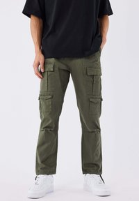 Persoon draagt olijfgroene cargobroek met meerdere zakken, een zwart T-shirt en witte sneakers, staand tegen een effen achtergrond.