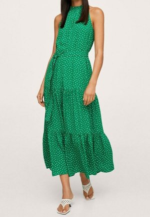 Grünes ärmelloses Kleid mit Neckholder, weißen Polka-Dots, tailliertem Bund, gestuftem Rock und seitlicher Schleifendetail. Kombiniert mit weißen Sandalen.