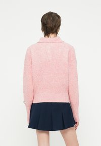 Rosa gestrickter Pullover mit Kragen, gerippter Textur und überschnittenen Schultern. Kombiniert mit einem navy-farbenen, plissierten Rock, der einen lässigen Schichtlook zeigt.