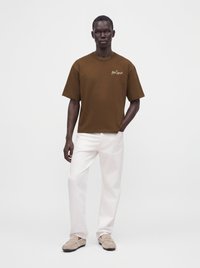 Άνδρας που φοράει ένα καφέ t-shirt Polo Sport, λευκό παντελόνι και μπεζ παπούτσια, στέκεται με το ένα χέρι στην τσέπη μπροστά σε ένα μονόχρωμο φόντο.