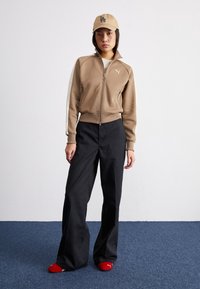 Femme portant une veste beige zippée, un pantalon large noir, une casquette beige avec le logo LA et des chaussettes rouges Puma, debout sur un tapis bleu contre un mur blanc.