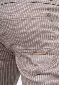 Gros plan sur un pantalon beige à fines rayures montrant la poche arrière avec une petite étiquette de marque en tissu et un détail de rivet métallique.