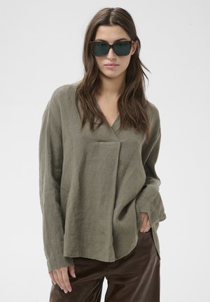 Žena so zelenými tmavými slnečnými okuliarmi, olivovo zeleným dlhorukávovým topom a tmavohnedými nohavicami, stojaca s jednou rukou v vrecku.